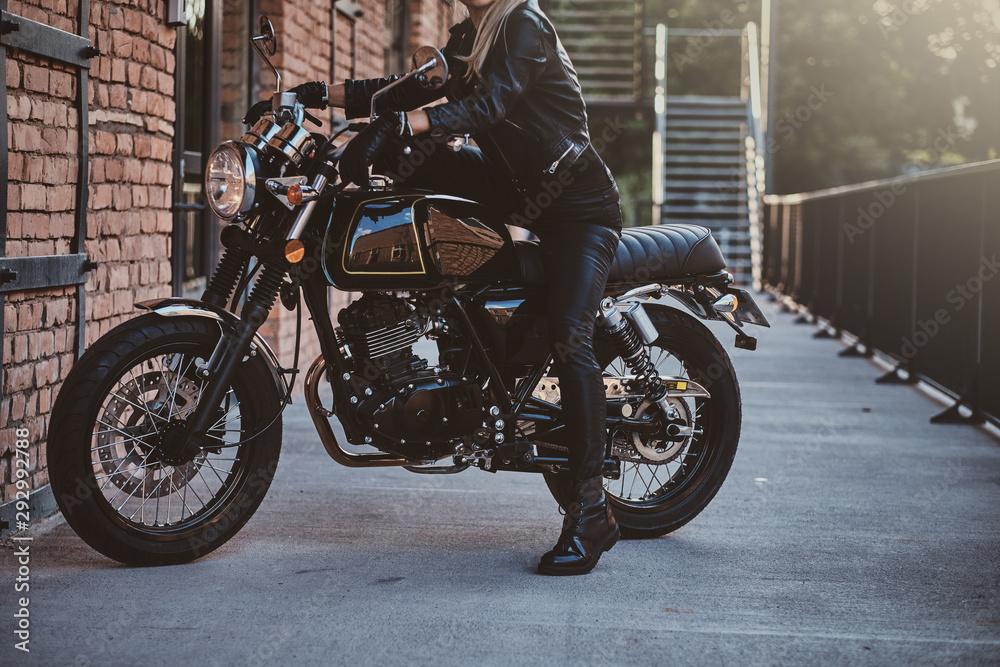 Stylish female biker is sitting at her black bike while posing for a photoshoot on the street.