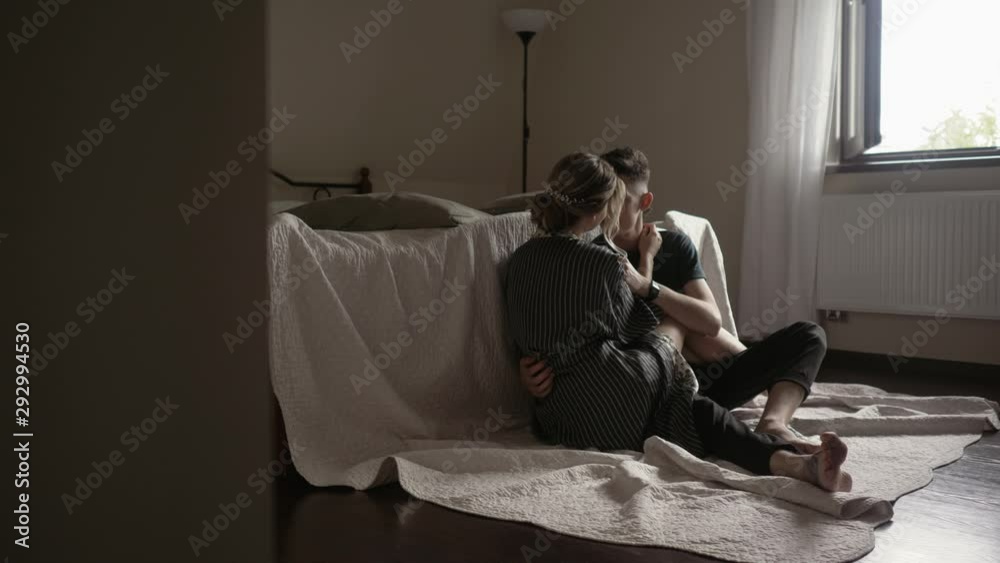 Young beautiful lovely couple is having a romantic time. Sitting on the floor by the bed embracing and smiling Young beautiful lovely couple is having a romantic time. Sitting on the floor by the bed embracing and smiling