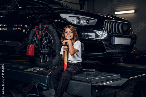 Beautiful small girl is sitting on the lifting platform with car and holding hammer.