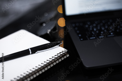 Close-up of pen and notebook on black desk of glass near laptop and sunglasses.