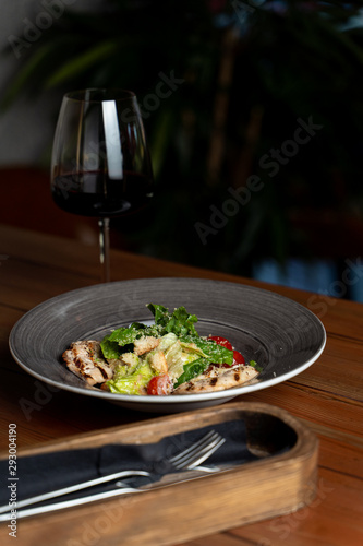 Side view of classic caesar salad with grilled chicken fillet, greens and cherry tomatoes with a glass of red wine