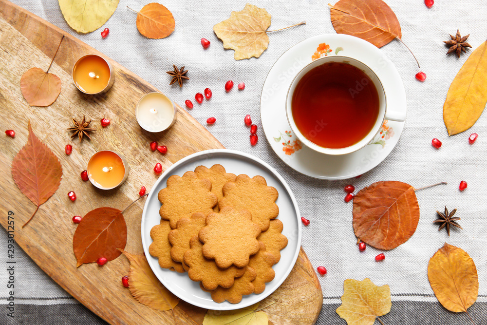 Fototapeta premium Top view of a cozy homemade tea party. Autumn composition. Cup of hot tea, Gingerbread cookie, burning candles, yellow fallen leaves, seeds pomegranate, anise on a linen tablecloth. Flat lay