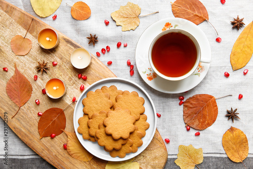 Top view of a cozy homemade tea party. Autumn composition. Cup of hot tea, Gingerbread cookie, burning candles, yellow fallen leaves, seeds pomegranate, anise on a linen tablecloth. Flat lay