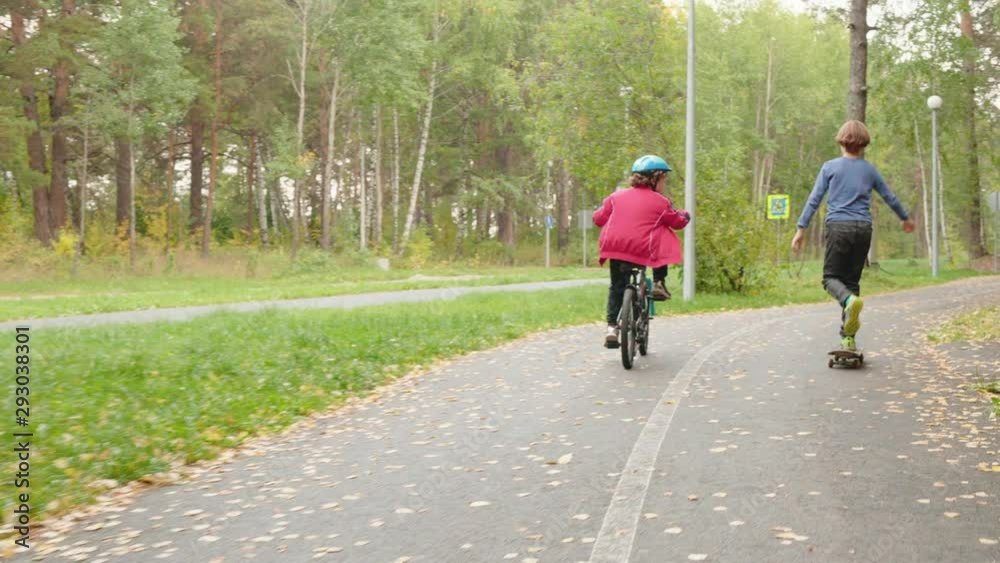 Children riding bicycle and skateboard in park. Back view of two preteen boys riding bike and longboard in beautiful autumn park. Leisure activity concept Children riding bicycle and skateboard in park. Back view of two preteen boys riding bike and longboard in beautiful autumn park. Leisure activity concept