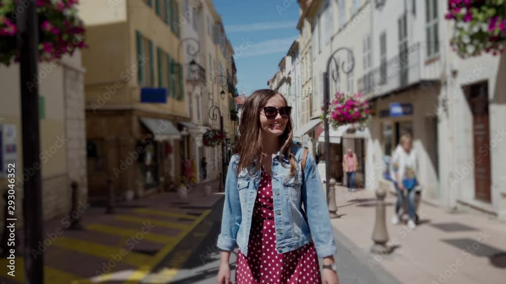 Young beautiful woman with sunglasses walking in the middle of the old town street on a sunny day. Female traveler wearing romantic red polka-dot dress. Blurred perspective view of narrow street with Young beautiful woman with sunglasses walking in the middle of the old town street on a sunny day. Female traveler wearing romantic red polka-dot dress. Blurred perspective view of narrow street with