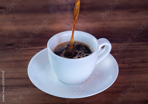 Coffee pouring in white cup, close up view. Coffee cup full of aromatic espresso. White cup and saucer on wooden table. Selective soft focus. Blurred background