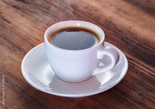 Close up view on smoking coffee in white cup. White coffee cup and saucer on brown table. Texture of wooden desk. Selective soft focus. Blurred background