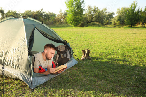 Young man in sleeping bag with book lying inside camping tent