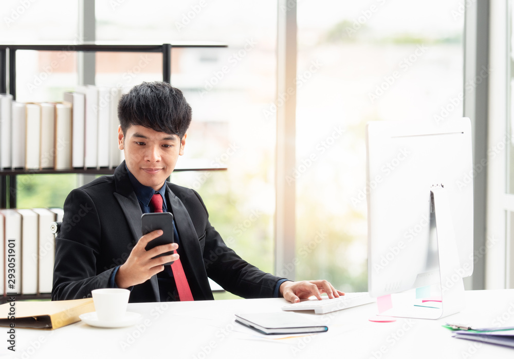 A young businessman with a confident suit sitting at a desk in a modern office. He is holding a phone to contact about the job while using a computer. Close up at the phone with copy space. A young businessman with a confident suit sitting at a desk in a modern office. He is holding a phone to contact about the job while using a computer. Close up at the phone with copy space.