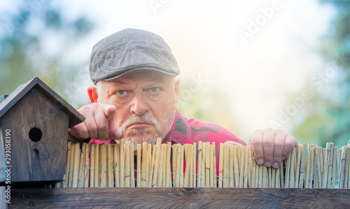 An elderly man in a hat looks angry and watches the neighborhood over a garden fence. Concept: Problems with the neighborhood. photo