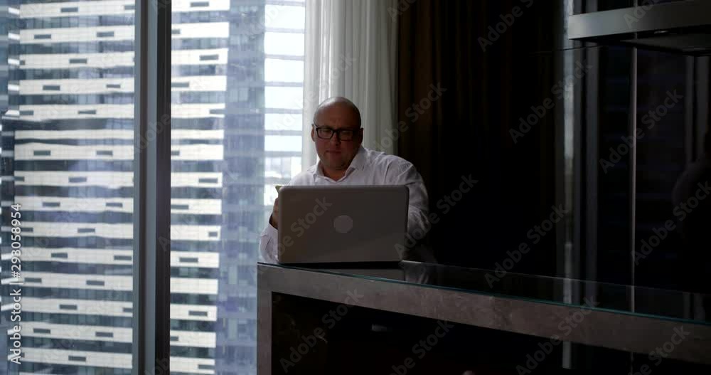 A bald businessman in glasses and a white shirt sits at a counter near a panoramic window in the business center, in front of him is a laptop, in his hands he holds a phone. A bald businessman in glasses and a white shirt sits at a counter near a panoramic window in the business center, in front of him is a laptop, in his hands he holds a phone.