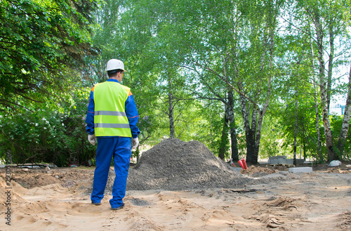 a worker in a blue uniform and a yellow vest, stands with his back in front of a pile of soil to repair the road