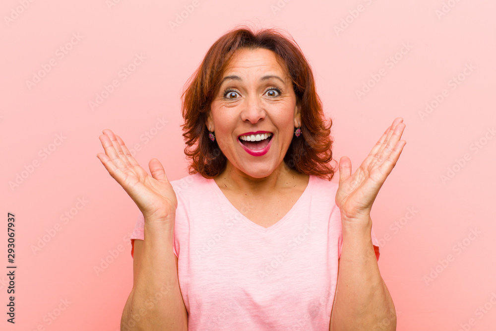 middle age woman looking happy and excited, shocked with an unexpected surprise with both hands open next to face against pink wall - obrazy, fototapety, plakaty 