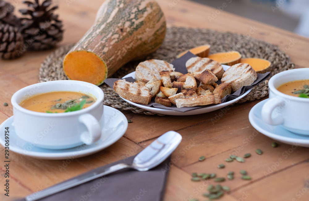 Close-up of two bowls of pumpkin cream soup. In the middle of the wooden table bread croutons.