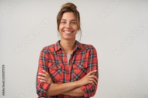 Studio portrait of young pretty dark haired female with bun hairstyle wearing casual clothes, posing over white background, looking happily to camera with crossed arms on chest