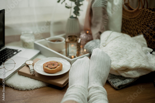 Toned photo. Autumn decor. Women's legs in knitted socks, a laptop, a mug of hot tea, a bun, candles. Cozy. Autumn.