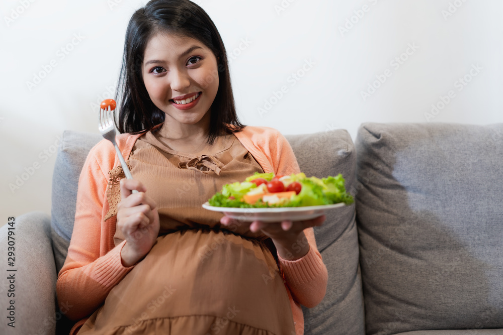 Happy pregnant Asian woman sitting and eating natural vegetable salad healthy food and sitting on sofa Happy pregnant Asian woman sitting and eating natural vegetable salad healthy food and sitting on sofa