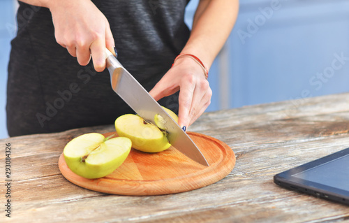 A young woman in a black t-shirt cuts green apple on a light wooden board in a kitchen.