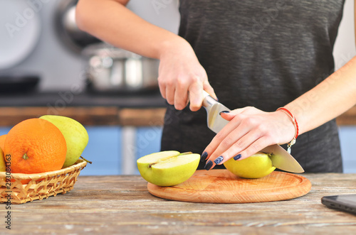 A young woman in a black t-shirt cuts green apple on a light wooden board in a kitchen.