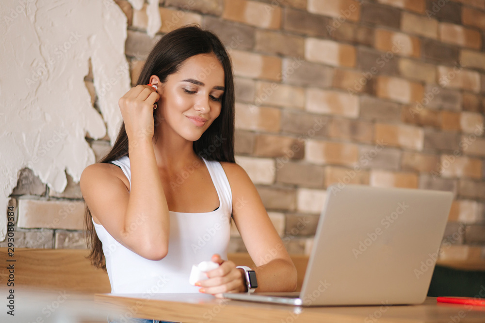 Attractive young girl use wirreless headphones and workin on her laptop. Woman in white t-shirt and denim. Student study in cafe Attractive young girl use wirreless headphones and workin on her laptop. Woman in white t-shirt and denim. Student study in cafe