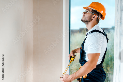 handsome repairman in goggles holding measuring tape and looking at wall near window