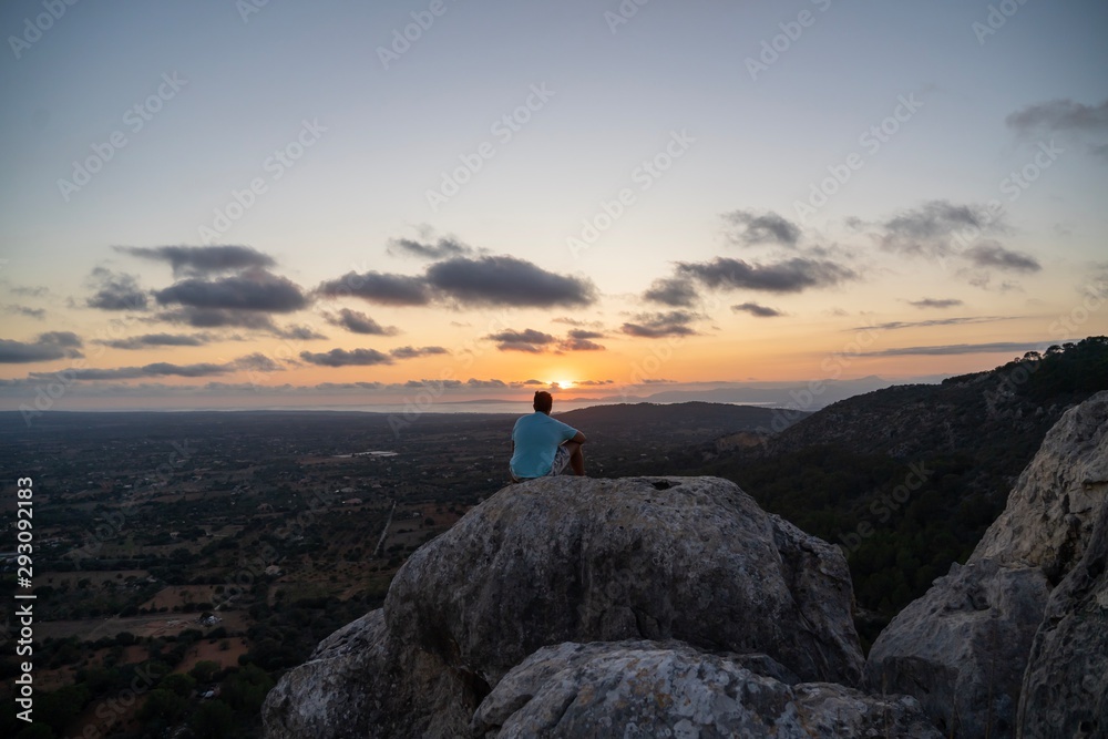 Young man with blue shirt sitting on a rock at the peak of a mountain while watching the sun setting out with some clouds in the sky and the rural lands Mallorca.