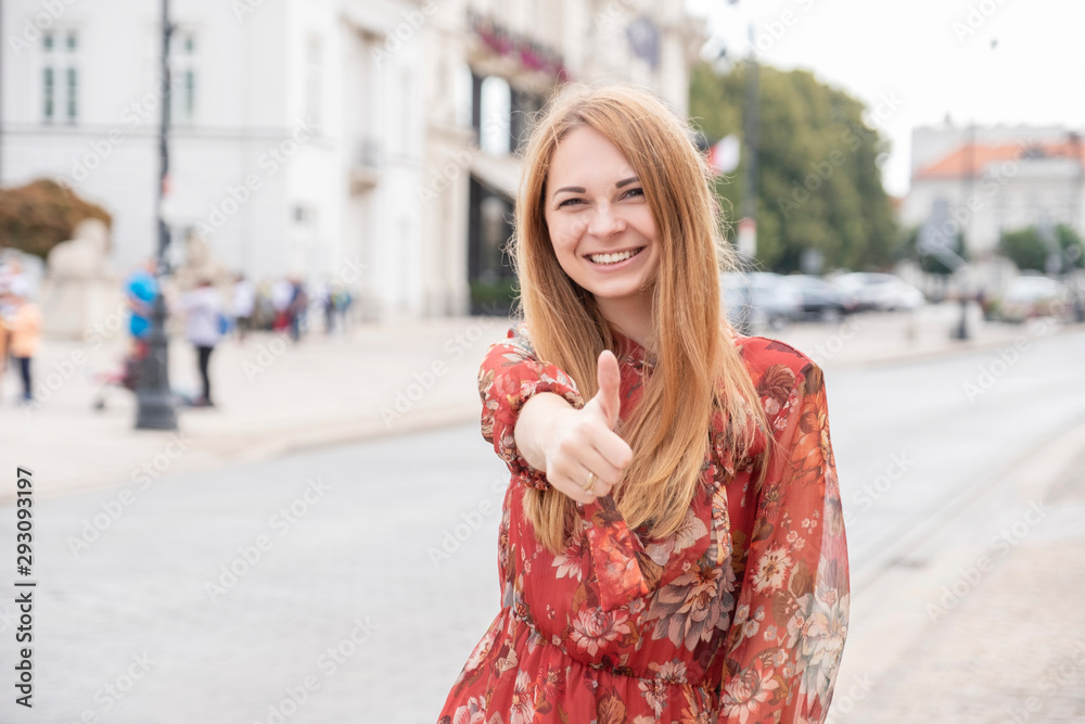Cute cheerful caucasian woman walking on european street. She wear stylish outfit and make thumb up with her hand Cute cheerful caucasian woman walking on european street. She wear stylish outfit and make thumb up with her hand