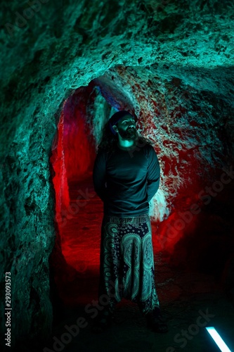 Young bearded caucasian man standing near a wall inside a narrow cave with blue and green lights. The model stays focused while a wall between the photographer and him stays unfocused.