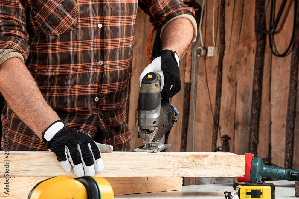 A man carpenter cuts a wooden beam using an electric jigsaw, male hands with an electric jigsaw closeup. Work with wood. A man carpenter cuts a wooden beam using an electric jigsaw, male hands with an electric jigsaw closeup. Work with wood.
