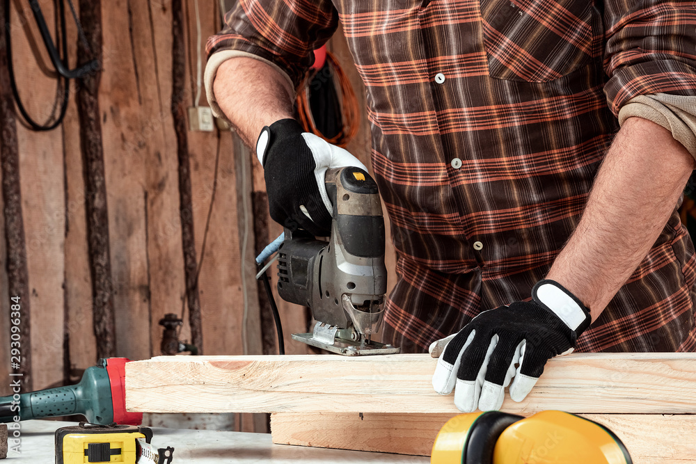 A man carpenter cuts a wooden beam using an electric jigsaw, male hands with an electric jigsaw closeup. Work with wood. A man carpenter cuts a wooden beam using an electric jigsaw, male hands with an electric jigsaw closeup. Work with wood.