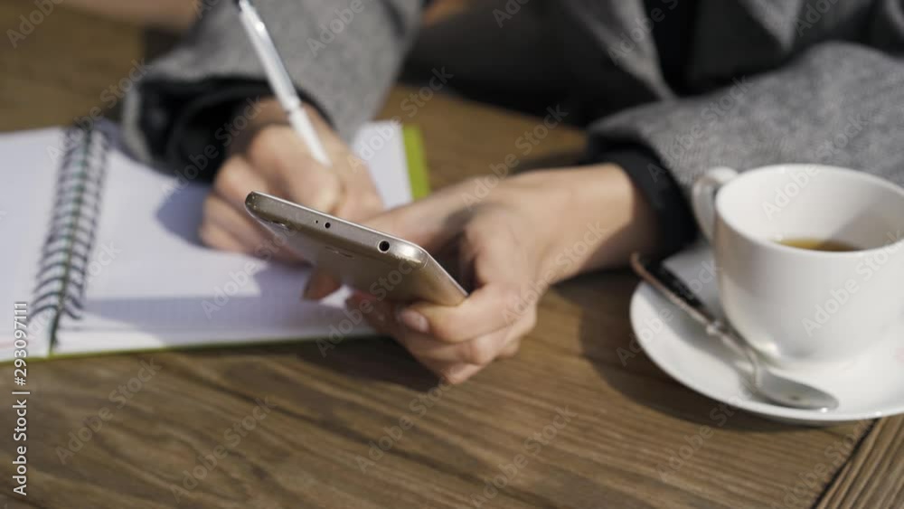Close-up of young caucasian woman holding cell phone while sitting at the table in the cafe. The girl writting in her notebook. Freelancer working. Autumn leisure indoors