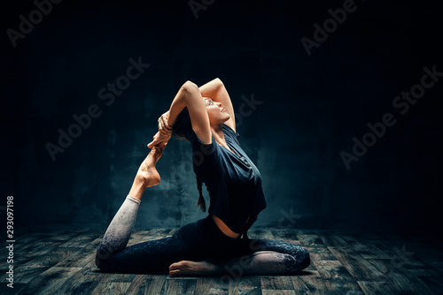 Young woman practicing yoga doing one legged king pigeon pose in dark room
