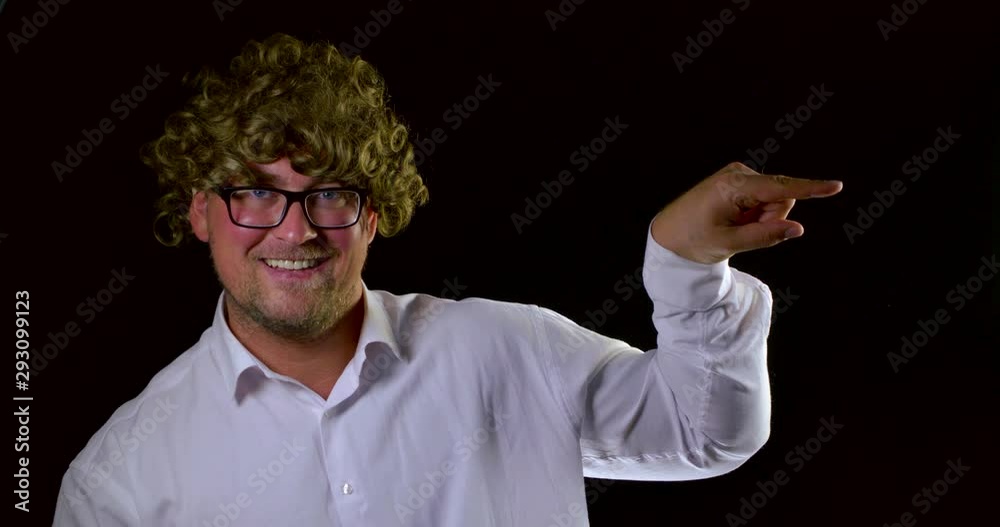 A close-up portrait of a curly-haired man in glasses and a white shirt, he is on a black background in the Studio, emoting, smiling and pointing his finger to the side. A close-up portrait of a curly-haired man in glasses and a white shirt, he is on a black background in the Studio, emoting, smiling and pointing his finger to the side.