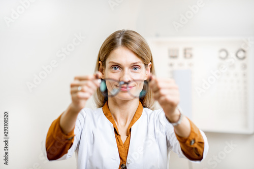 Ophthalmologist offering eyeglasses putting them forward to the camera, standing in front of eye chart in the medical office