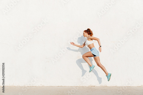 Image of redhead young woman running along white wall while doing workout in morning photo