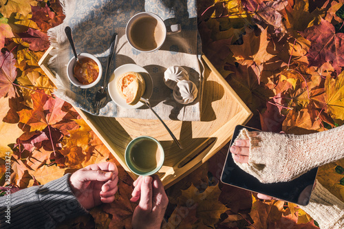Couple of young people are taking a picture on a tablet of morning breakfast on a wooden tray in the autumn park with colorful maple leaves. Aerial view