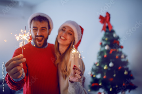 Happy caucasian couple with santa hats on heads holding sprinklers while hugging and standing in living room. In background is christmas tree. Selective focus on sprinklers.