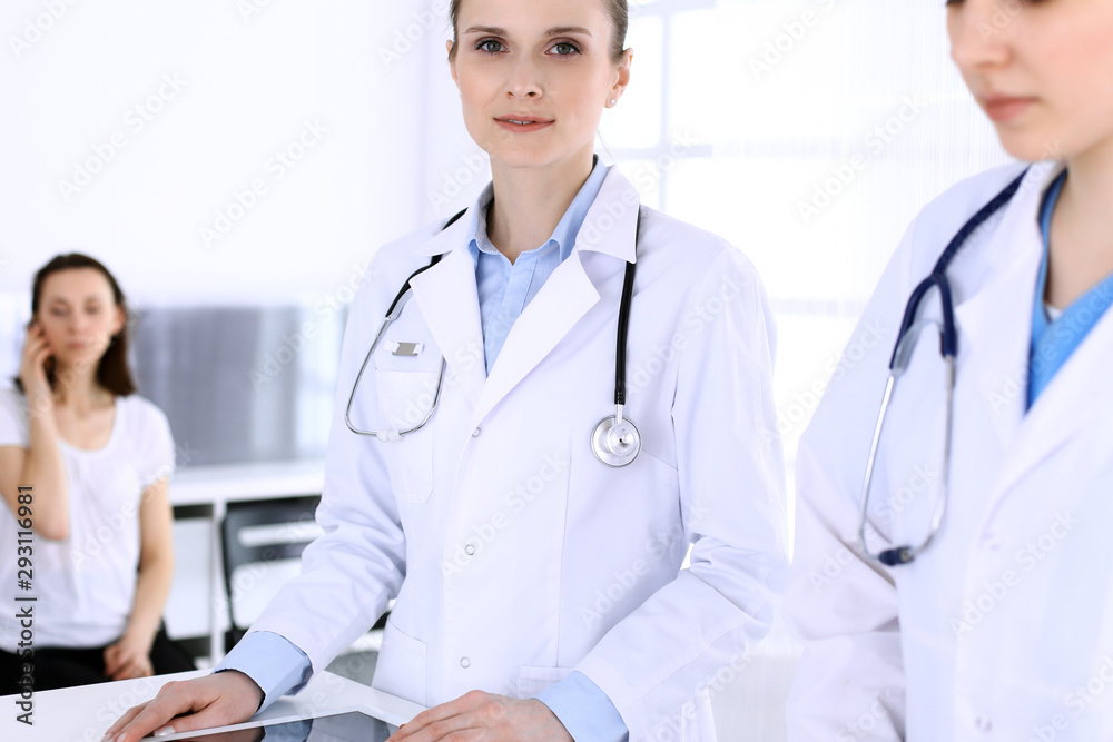 Group of doctors at work in hospital with patient in a queue at the background. Physician filling up medical documents or prescription while standing at reception desk. Data in medicine and health Group of doctors at work in hospital with patient in a queue at the background. Physician filling up medical documents or prescription while standing at reception desk. Data in medicine and health