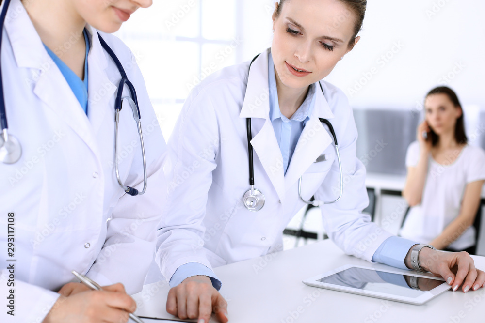 Group of doctors at work in hospital with patient in a queue at the background. Physician filling up medical documents or prescription while standing at reception desk. Data in medicine and health Group of doctors at work in hospital with patient in a queue at the background. Physician filling up medical documents or prescription while standing at reception desk. Data in medicine and health