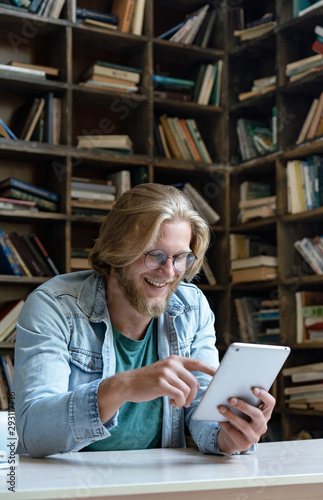 Happy male student teacher holding using digital tablet computer looking at screen reading e book learning easy internet course study online in app concept with device sit at library desk, vertical photo