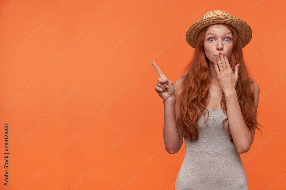Dazed attractive young lady with wavy foxy hair posing over orange background in casual clothes, raising hand and pointing aside with index finger, keeping palm on her mouth and raising eyebrows Dazed attractive young lady with wavy foxy hair posing over orange background in casual clothes, raising hand and pointing aside with index finger, keeping palm on her mouth and raising eyebrows