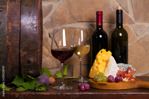 Fototapeta Naklejka Na Ścianę i Meble -  Red and white wine with  grapes beside old cask in wine cellar. Glasses and bottles of wine with cheese, jamon, figs and nuts