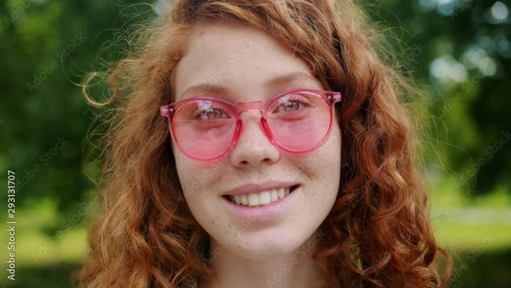 Close-up portrait of pretty redhead girl with freckles wearing sunglasses in park on summer day smiling looking at camera. Beautiful people and nature concept. Close-up portrait of pretty redhead girl with freckles wearing sunglasses in park on summer day smiling looking at camera. Beautiful people and nature concept.