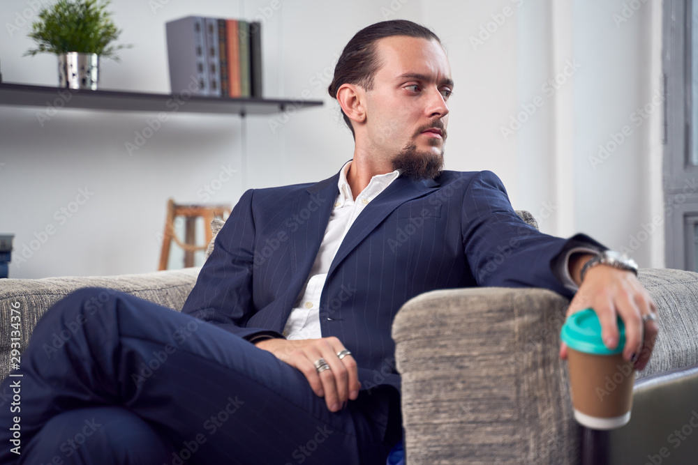 Photo of young brunett with glass of coffee in his hands sitting on sofa Photo of young brunett with glass of coffee in his hands sitting on sofa