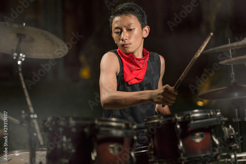  Handsome and cool Asian American teenager playing drum set at home garden emulating night music show as hobby in teenage love for music