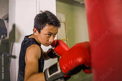 young handsome and fierce teenager boy doing fight workout punching heavy bag looking cool and badass at fitness club training hard using gloves © TheVisualsYouNeed