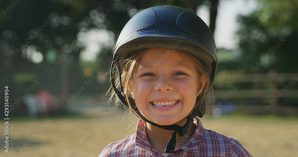 Authentic shot of a little girl with a helmet smiling in the camera at riding stable on a sunny day. Authentic shot of a little girl with a helmet smiling in the camera at riding stable on a sunny day.