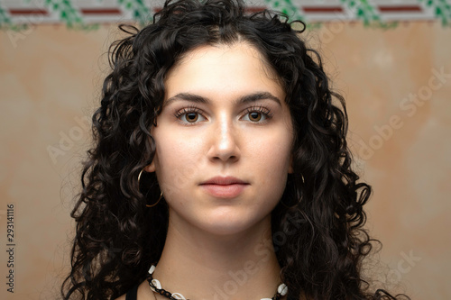 A closeup portrait of a beautiful young girl in her early twenties, with soft facial features and minimal makeup, luscious dark curly hair is seen against a neutral background.