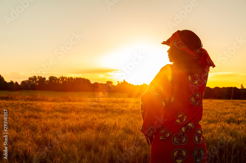 Leinwand Poster African woman in traditional clothes standing in a field of crops at sunset or s
