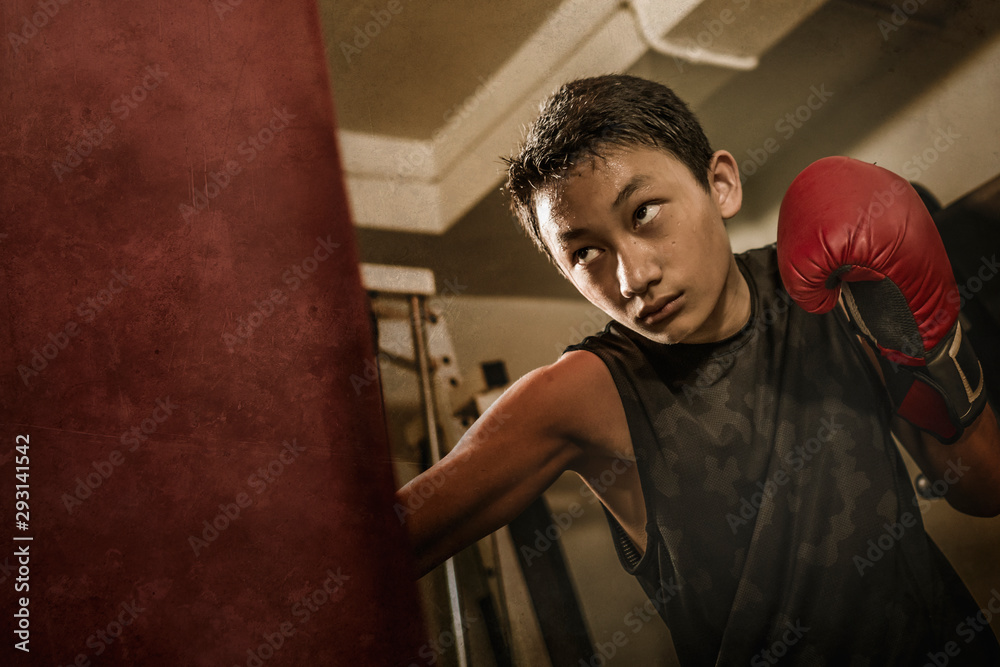 young handsome and fierce teenager boy doing fight workout punching heavy bag looking cool and badass at fitness club training hard using gloves